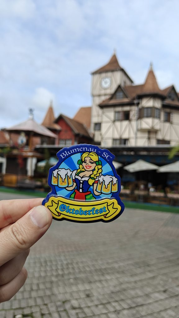 Ímã de geladeira da Oktoberfest de Blumenau (SC) com ilustração de mulher em traje típico alemão segurando canecas de chope, fotografado em frente à Vila Germânica.